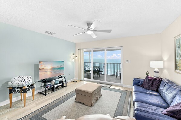 A living room with a television, a sofa, a coffee table, and a view of the ocean through a sliding glass door.
