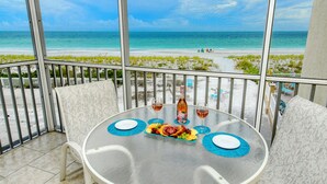 Mesa redonda com tampo de vidro, duas taças de vinho, uma garrafa e um prato de comida numa varanda com vista para a praia.