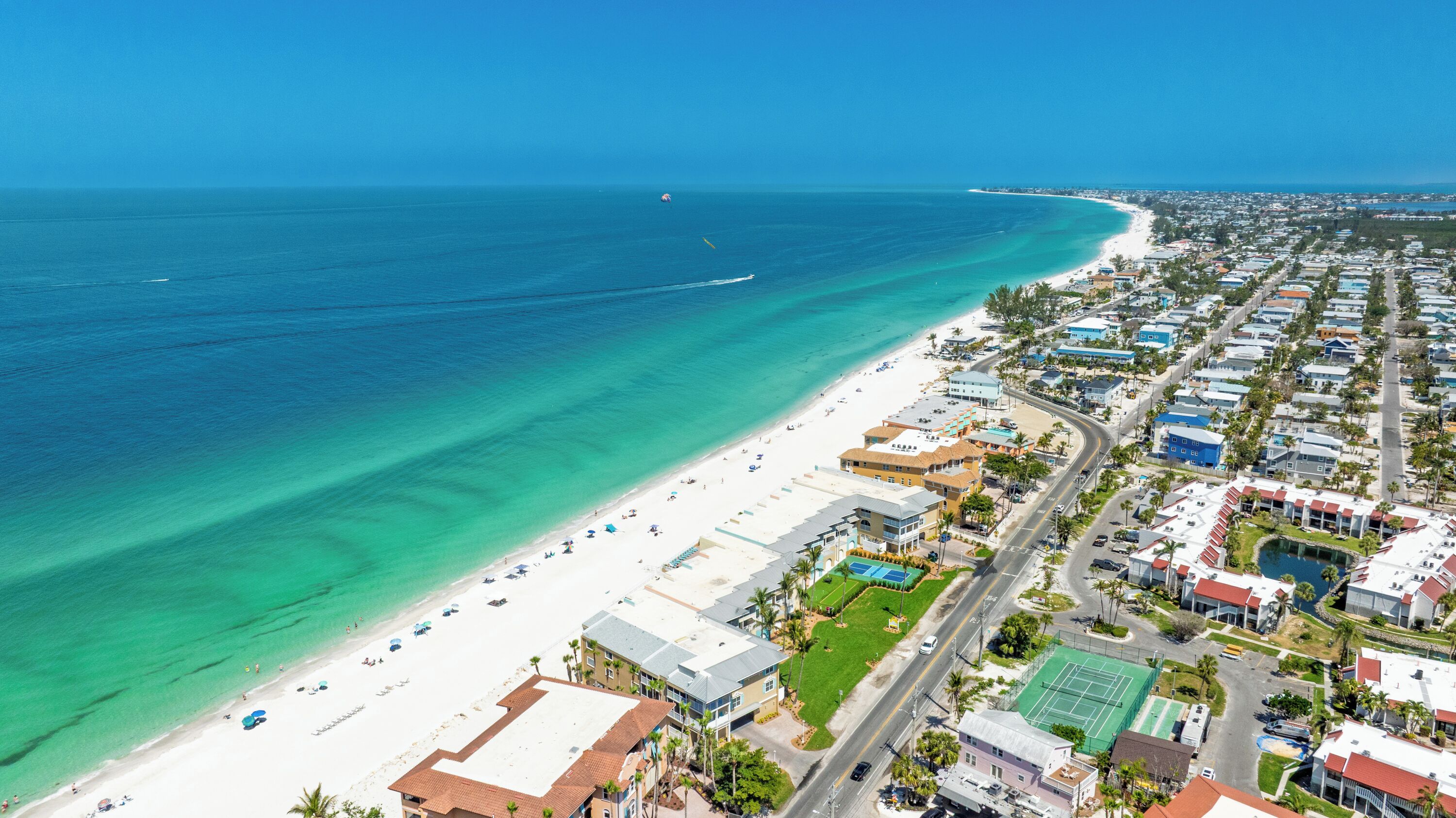 Vue aérienne d’une ville côtière avec une longue plage de sable, des eaux d’un bleu limpide et un mélange de bâtiments résidentiels et commerciaux.