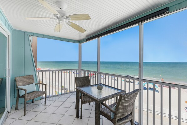 A covered balcony with wicker furniture, a table, and a view of the beach and ocean.