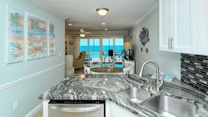 A kitchen with a granite countertop, stainless steel sink, and a dishwasher. There is a dining area with a table and chairs, and a view of the ocean through large windows. - Beautiful beachfront views!  Direct beach access! (Bradenton Beach)