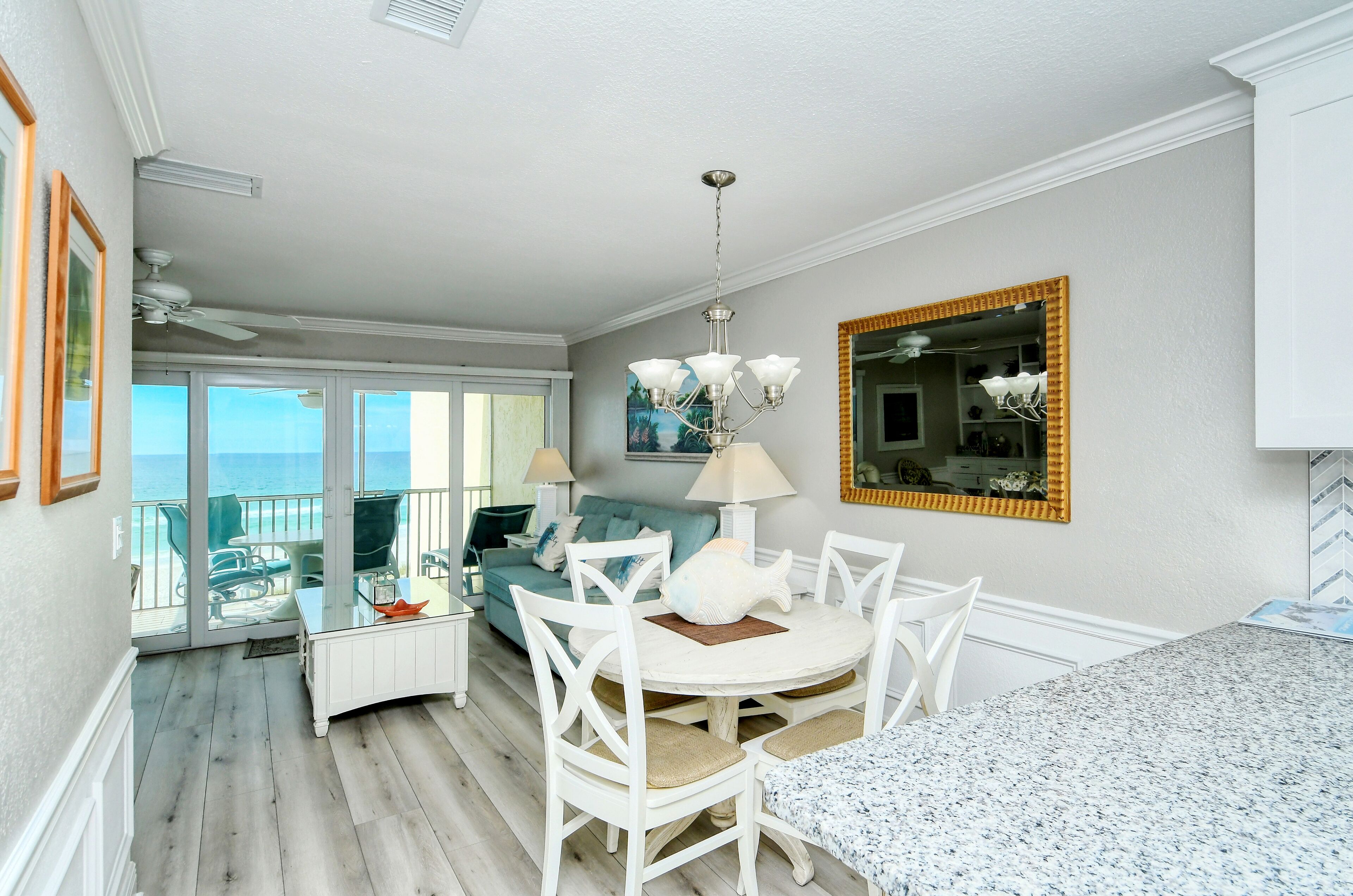 A dining area with a round table, white chairs, and a chandelier. There is a balcony with a view of the ocean.