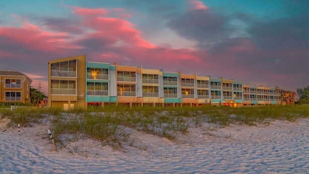 A beachfront hotel with multiple balconies, a sandy beach in front, and a colorful sunset sky. - Peak a boo Gulf of Mexico balcony views! Direct beach access! Summer and fall dates available! (Bradenton Beach)