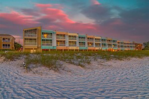 A beachfront hotel with multiple balconies, a sandy beach in front, and a colorful sunset sky.