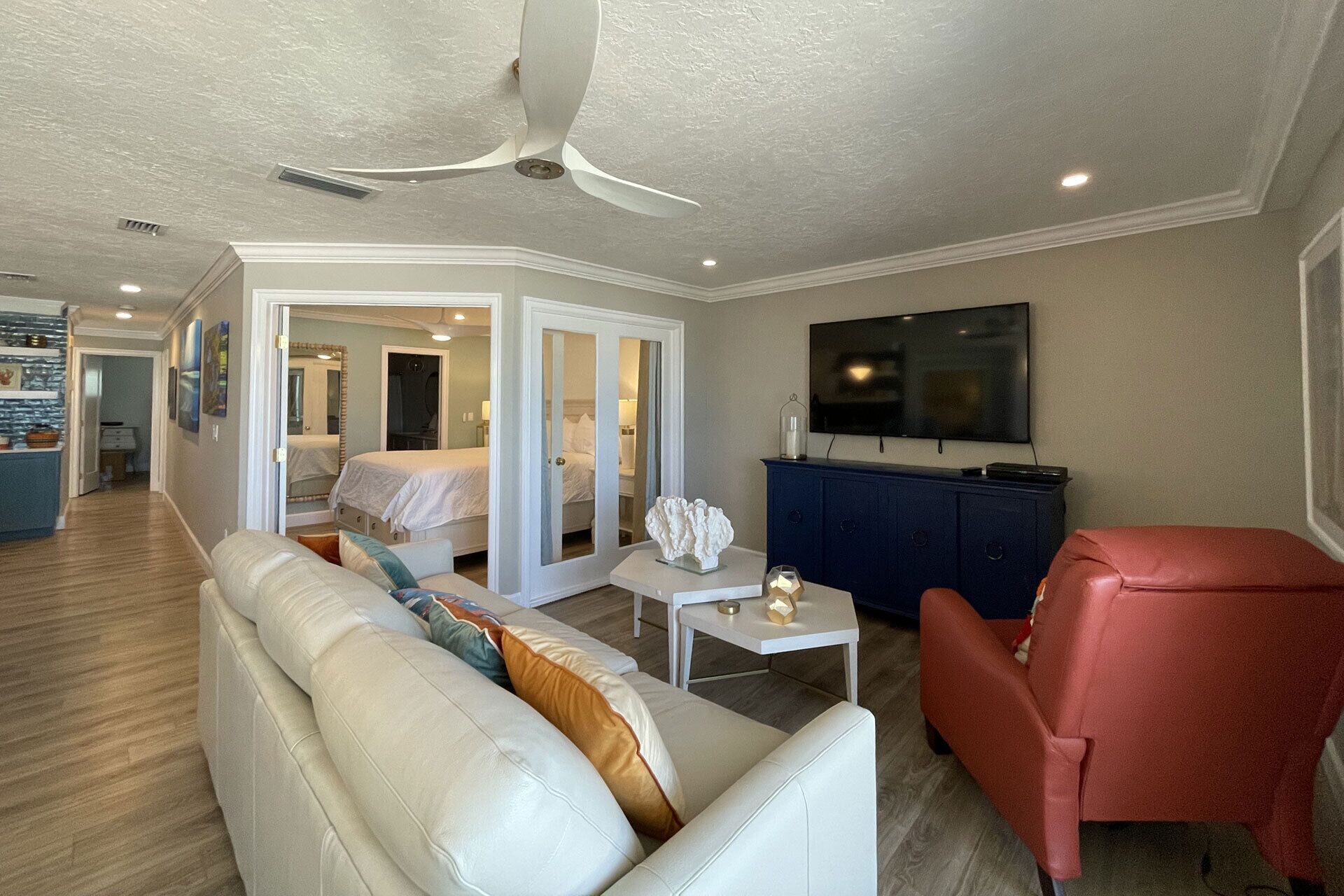 Habitación de hotel con sofá blanco, sillón rojo, mesita blanca pequeña, mueble azul y televisor en la pared.
