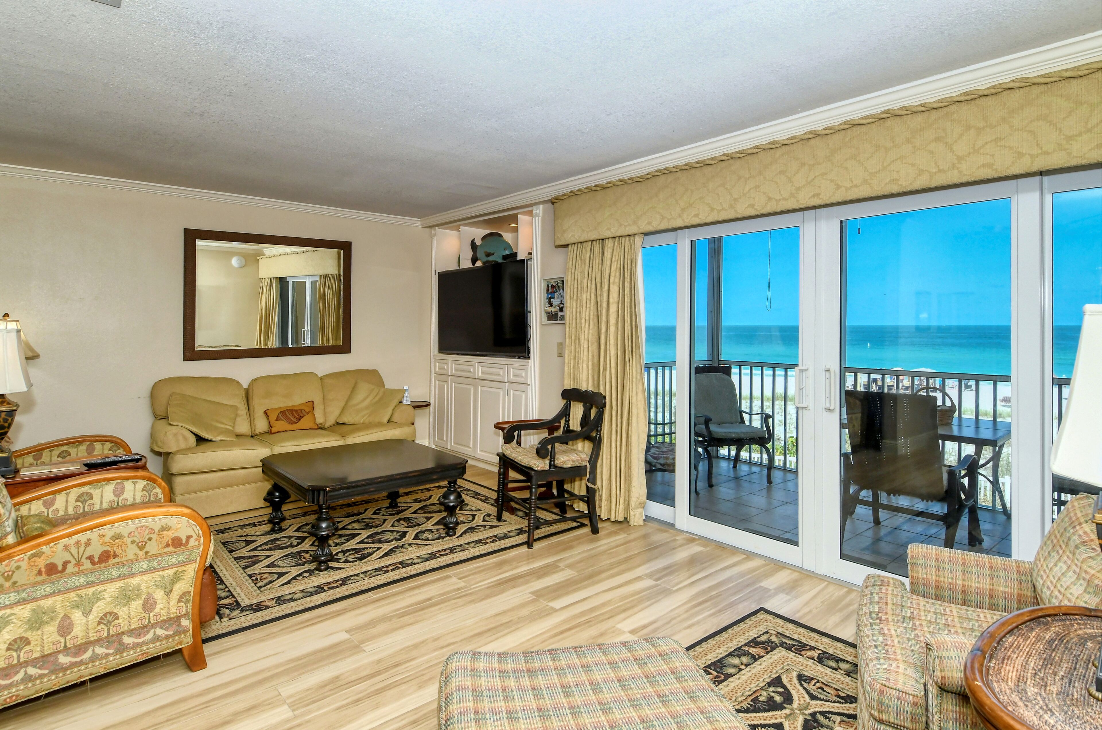 A living room with a sofa, armchairs, a coffee table, and a TV. There is a large window with a view of the ocean.