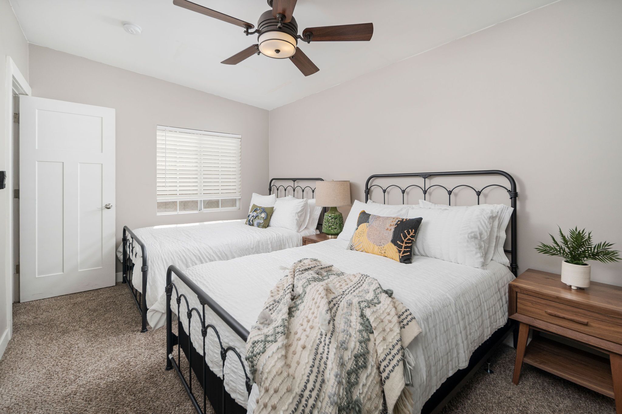 A bedroom with two beds, a ceiling fan, a window with blinds, and a bedside table with a plant.