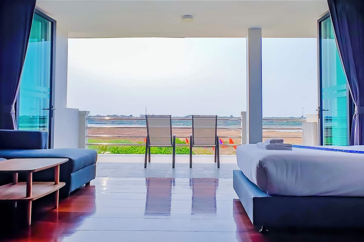 A modern hotel room with a large bed, a sofa, and a small table. There is a balcony with a view of the beach and the ocean.