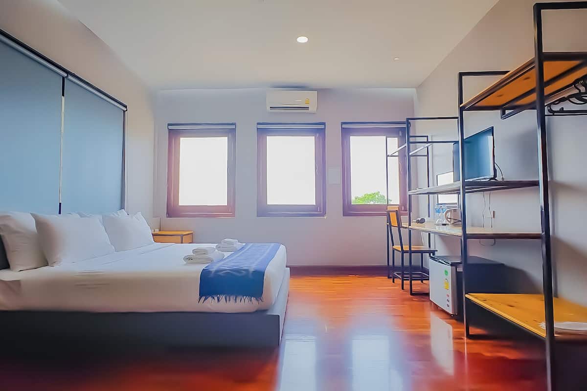 A bedroom with a bed, a desk, and shelving. There are three windows and an air conditioning unit.
