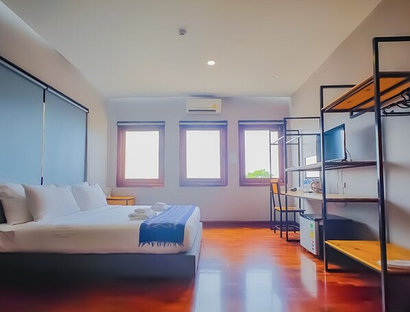 A bedroom with a bed, a desk, and shelving. There are three windows and an air conditioning unit.
