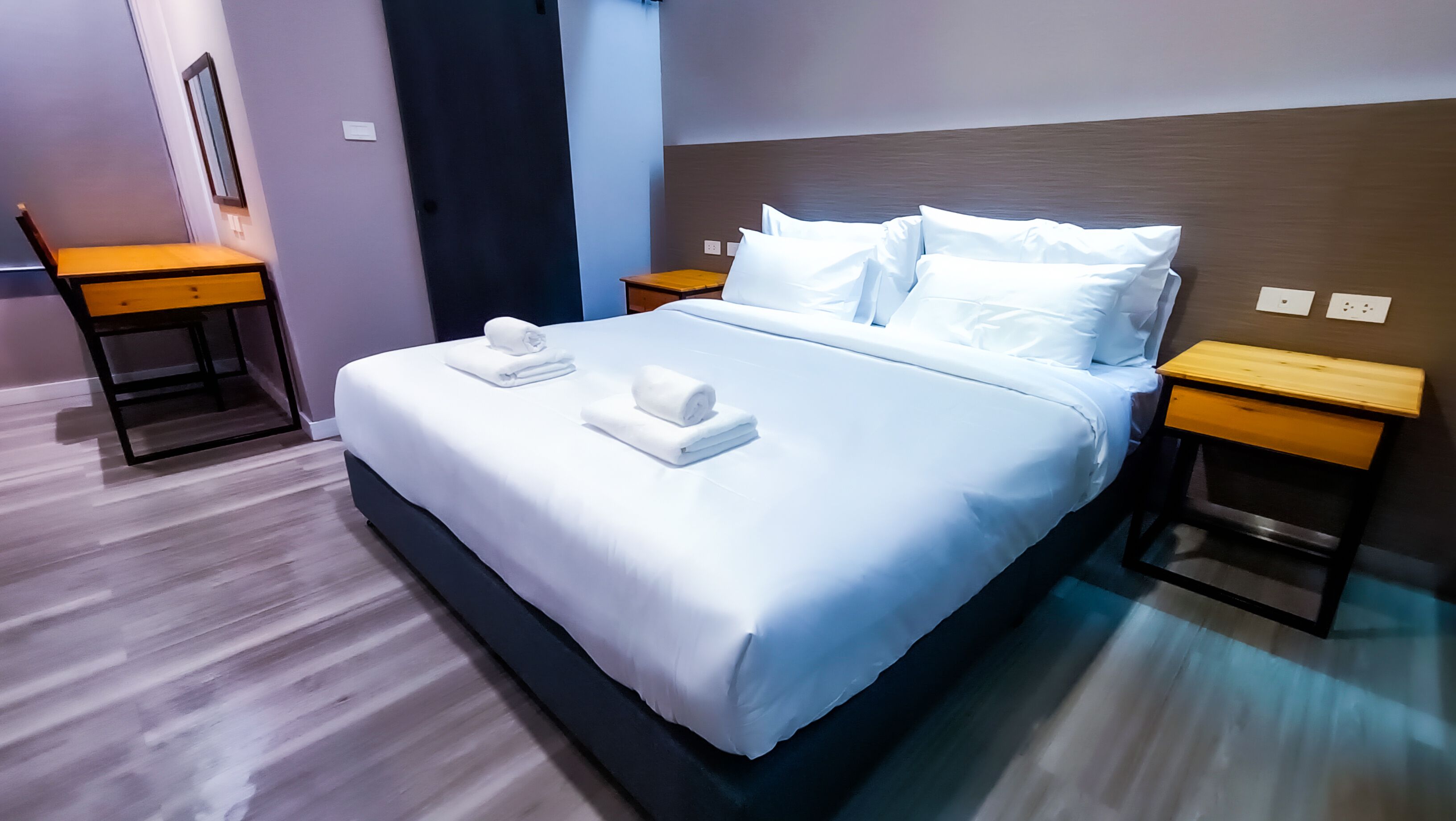 A neatly made bed with white linens and two rolled towels, wooden nightstands, and a mirror on the wall.