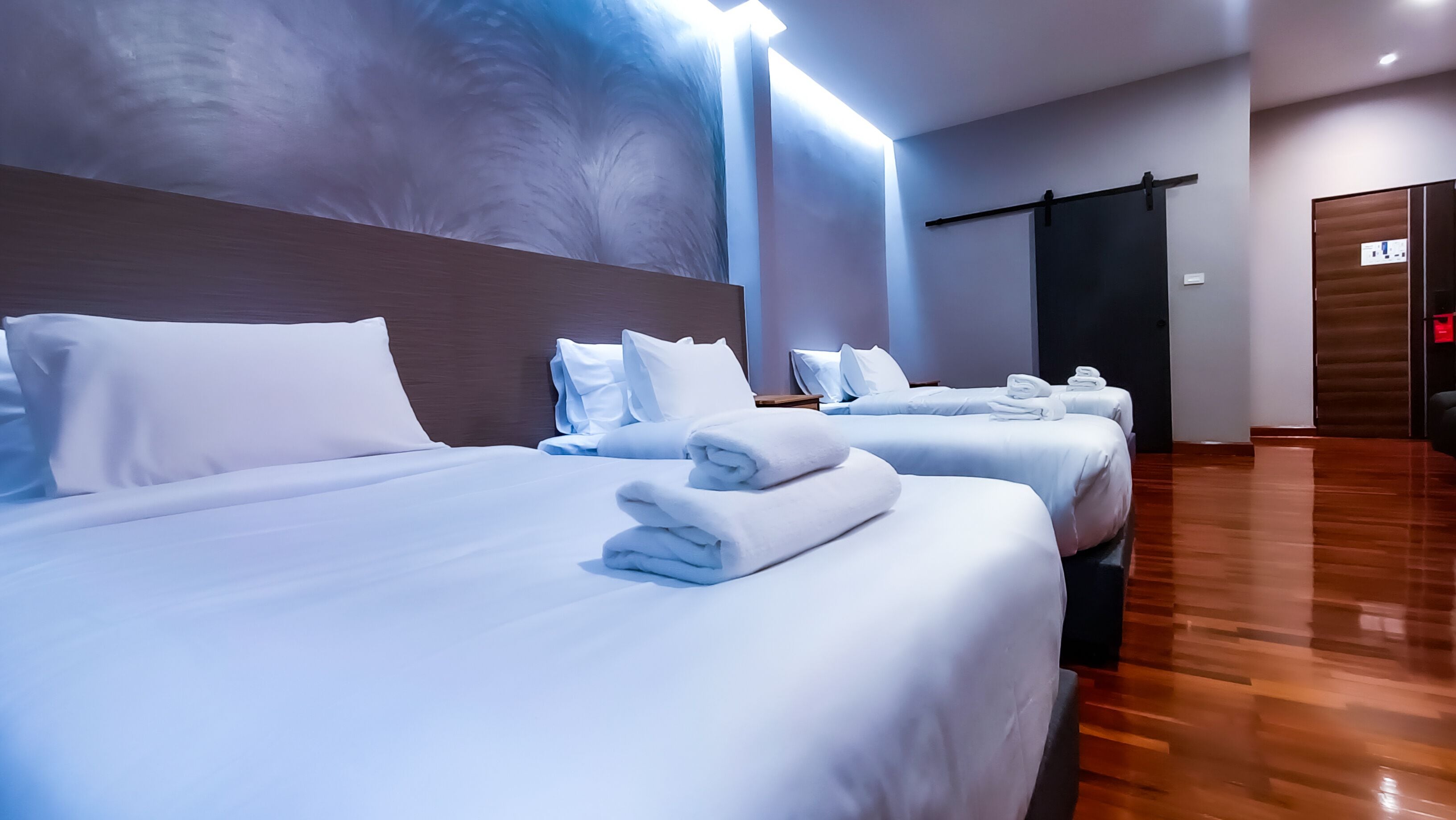 Ett hotellrum med tre sängar som är prydligt bäddade, alla med vita sängkläder och handdukar. Rummet har en modern design med ett trägolv och en stor, väggmonterad lampa.