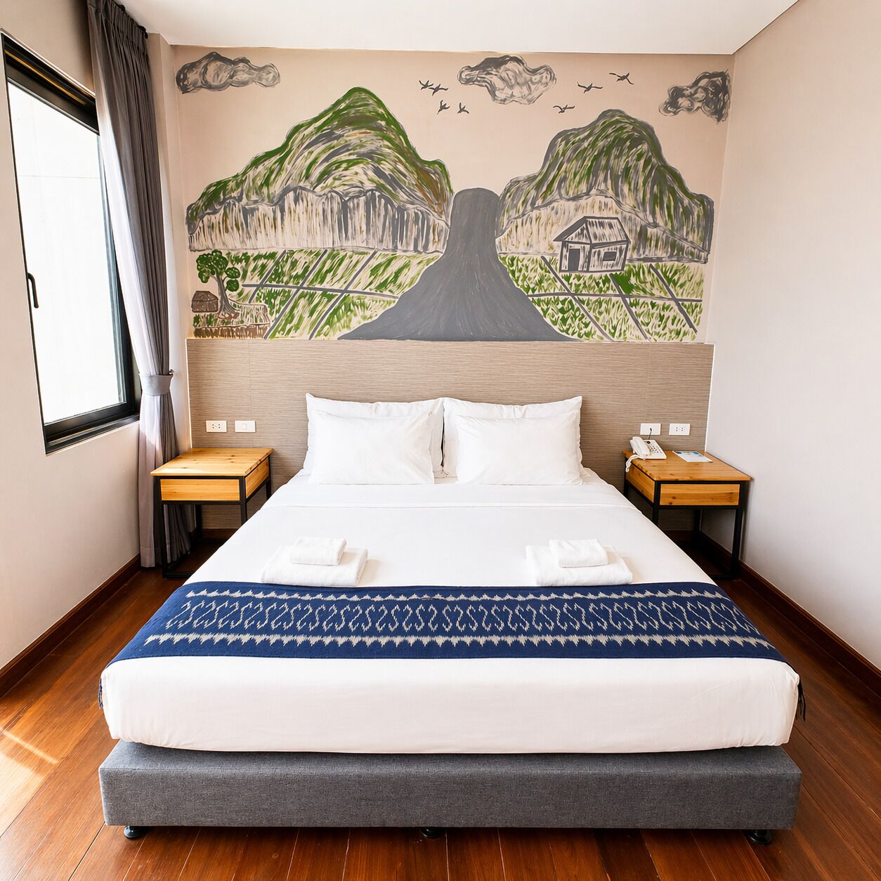 A hotel room with a bed, two bedside tables, a mural on the wall, and a window with curtains.