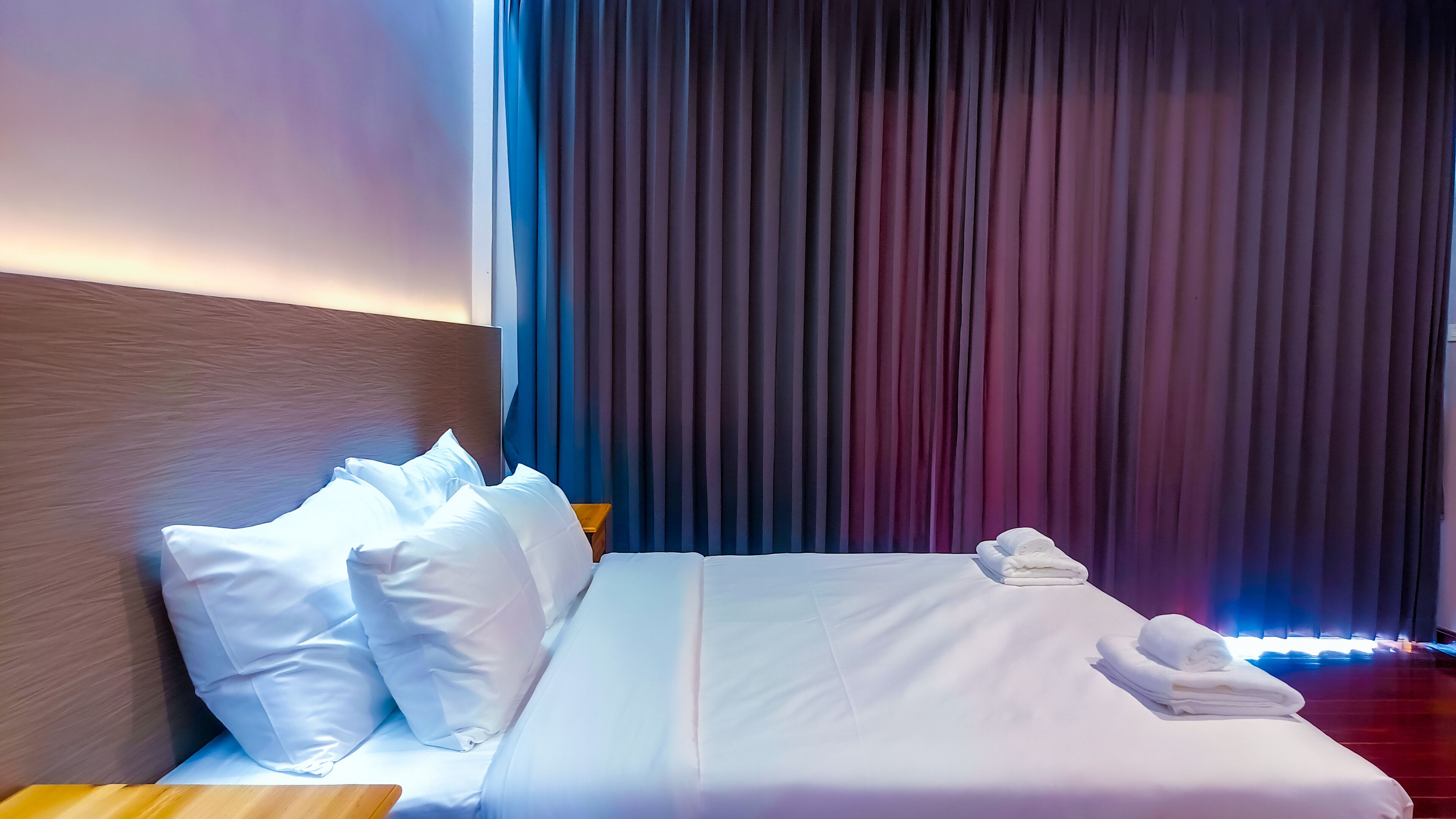 A neatly made bed with white linens and pillows, a wooden headboard, and a stack of folded towels on the foot of the bed.