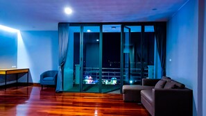 A modern living room with a wooden floor, a sofa, a chair, and a desk. There is a large sliding glass door leading to an outdoor balcony with a view of the city at night. - The Riva Vientiane Hotel (Vientiane)