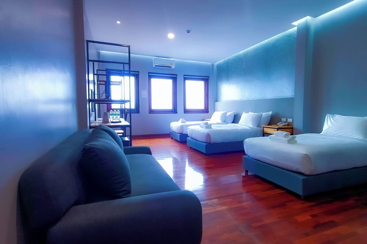 A modern hotel room with a sofa, two beds, a desk, and a view of the outdoors.