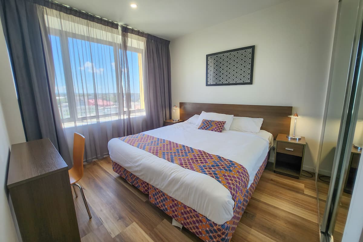 A hotel room with a large bed, a desk, and a chair. There is a window with curtains, a bedside table with a lamp, and a wall-mounted artwork.