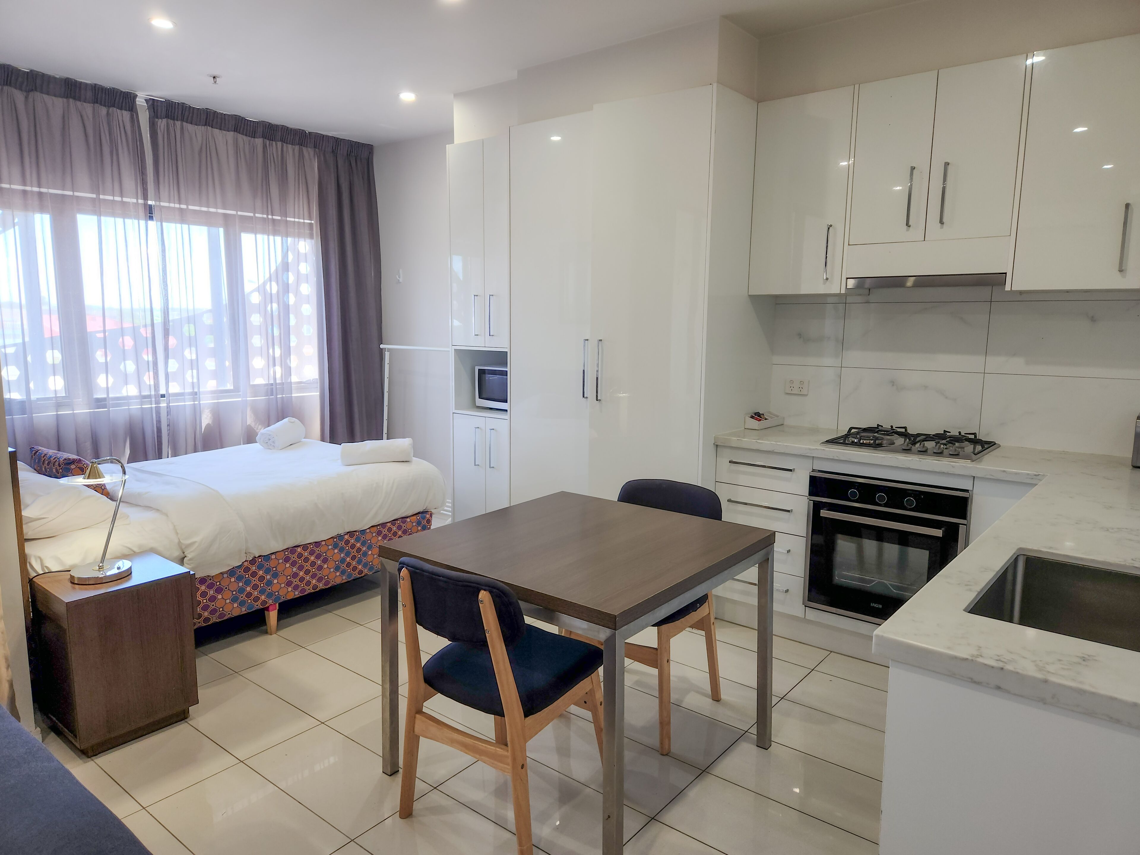 A modern hotel room with a bed, a desk, a chair, a TV, a kitchen area with a stove and oven, and a window with curtains.