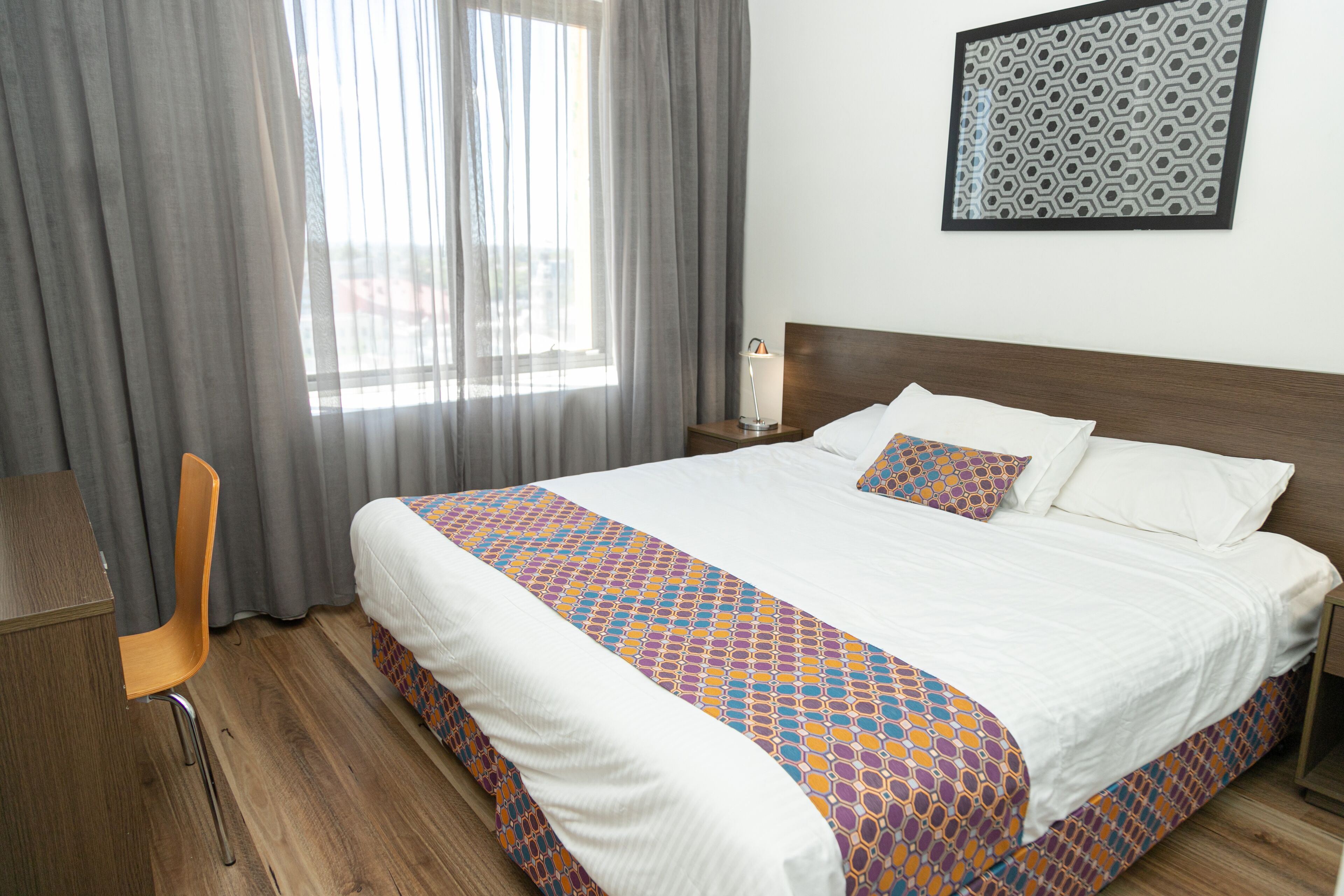 Una habitación de hotel con una cama grande, un escritorio de madera, una silla y un ventanal con cortinas.