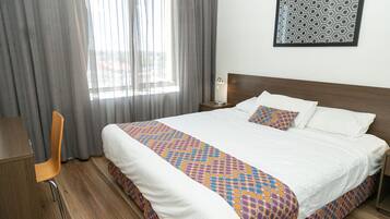 Una habitación de hotel con una cama grande, un escritorio de madera, una silla y un ventanal con cortinas.
