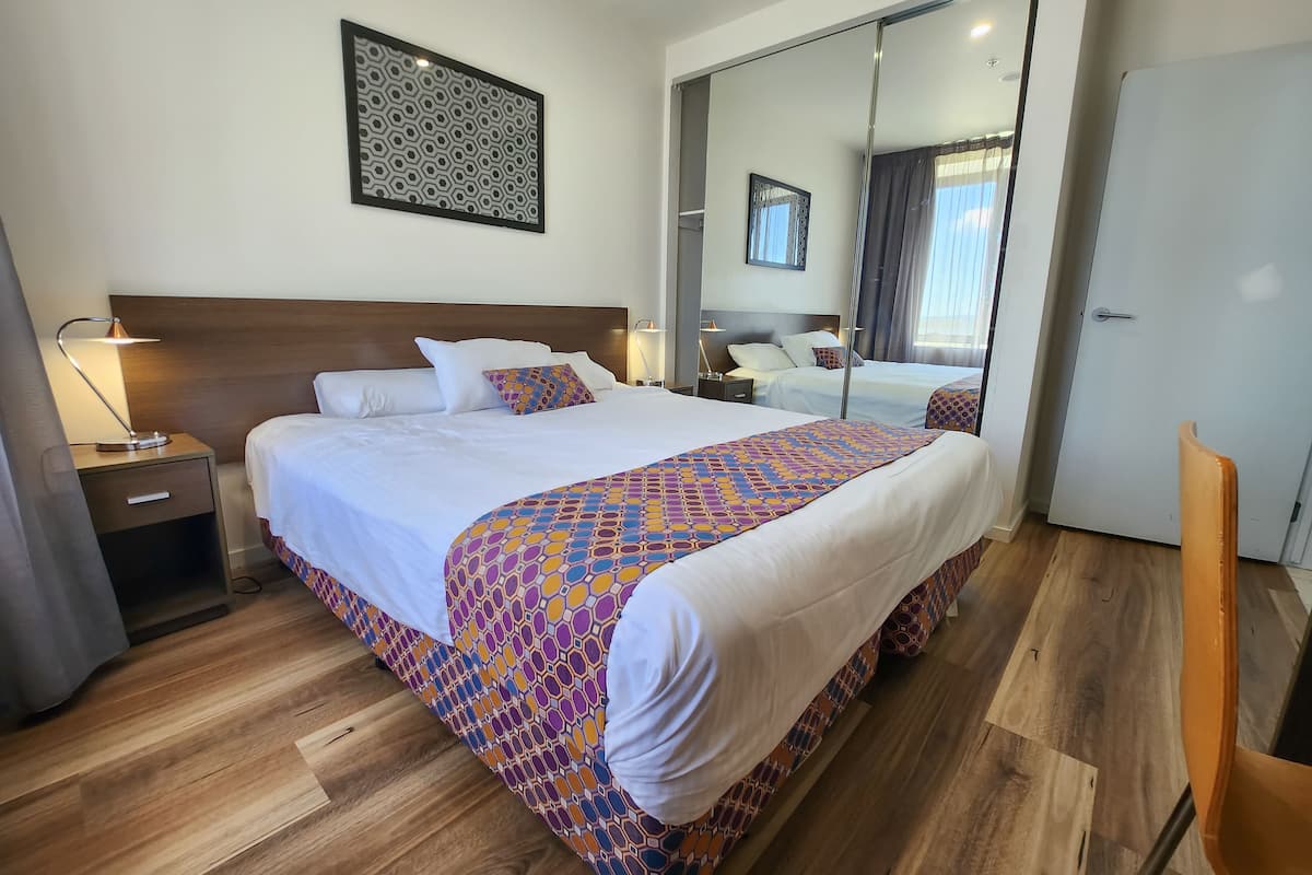 A hotel room with a large bed, a wooden headboard, a bedside table with a lamp, a mirror, and a door.