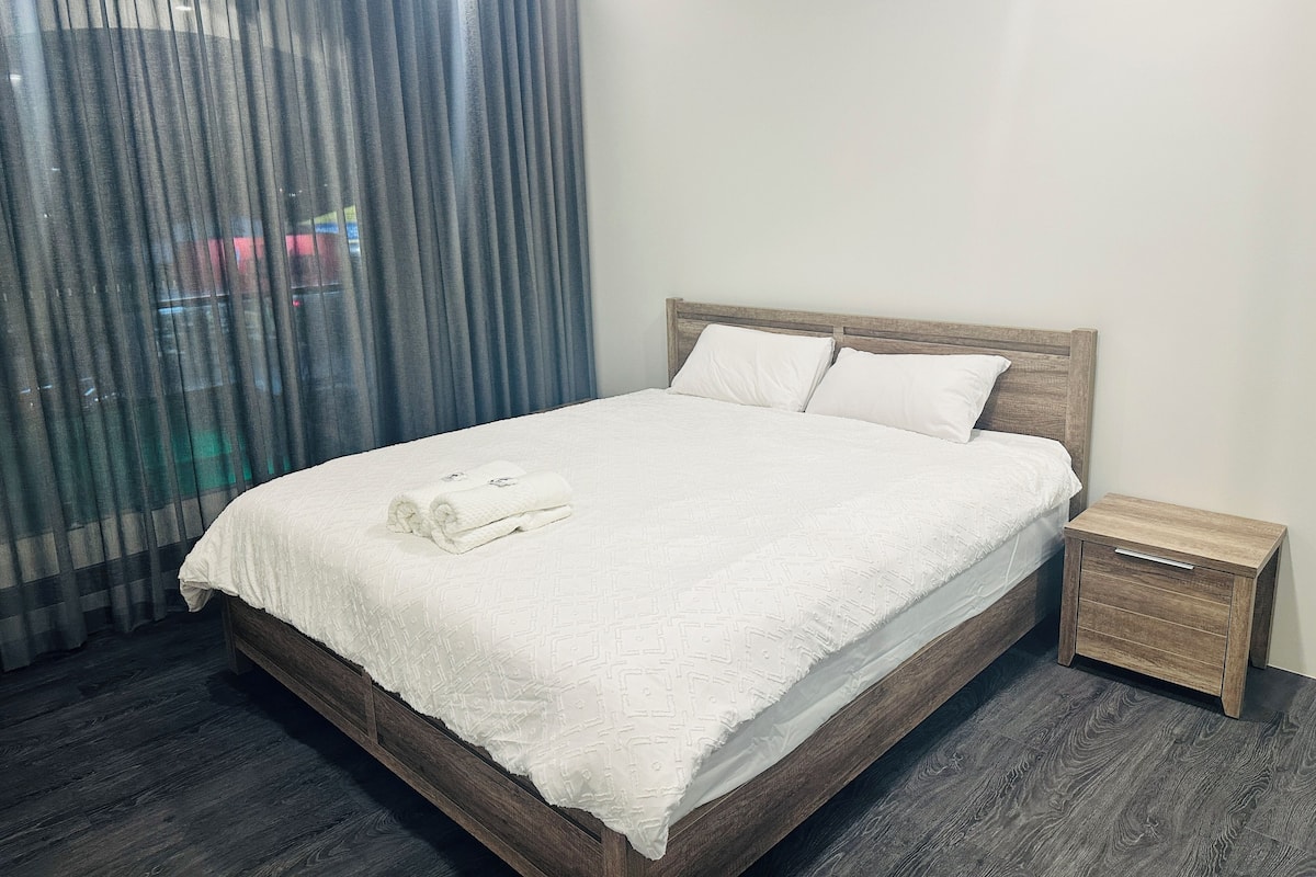 A neatly made bed with white linens and pillows, a wooden headboard, and a small wooden nightstand with a drawer.