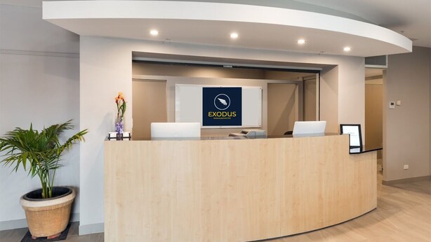 A reception desk with a logo, a potted plant, and a laptop. - Exodus Dandenong Apartment Hotel (Dandenong)