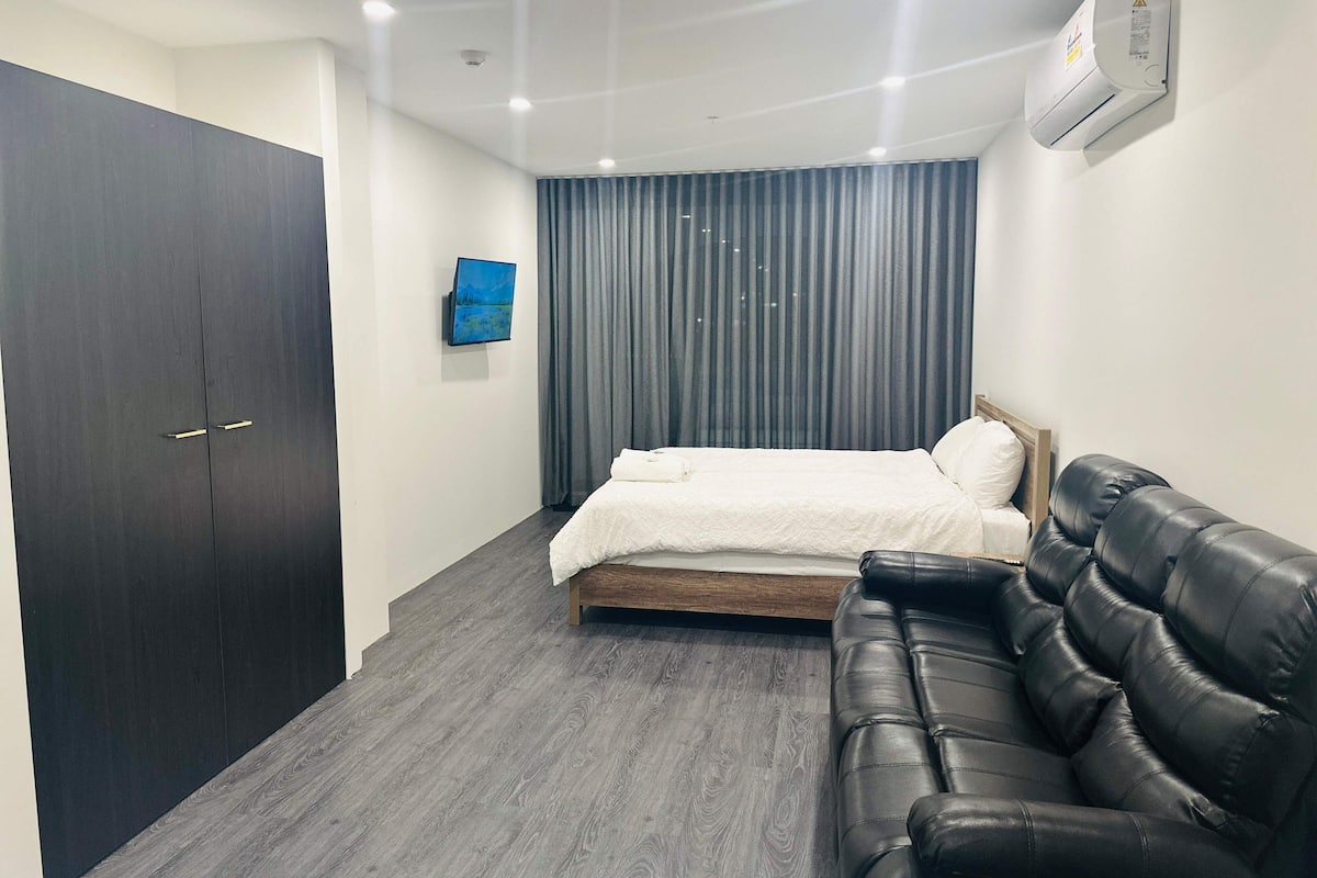 A modern bedroom with a bed, a dark wardrobe, a leather sofa, a mounted TV, and an air conditioning unit.