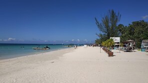 A beach with a sandy shore, a few boats, and a row of beachside structures with umbrellas and lounge chairs.