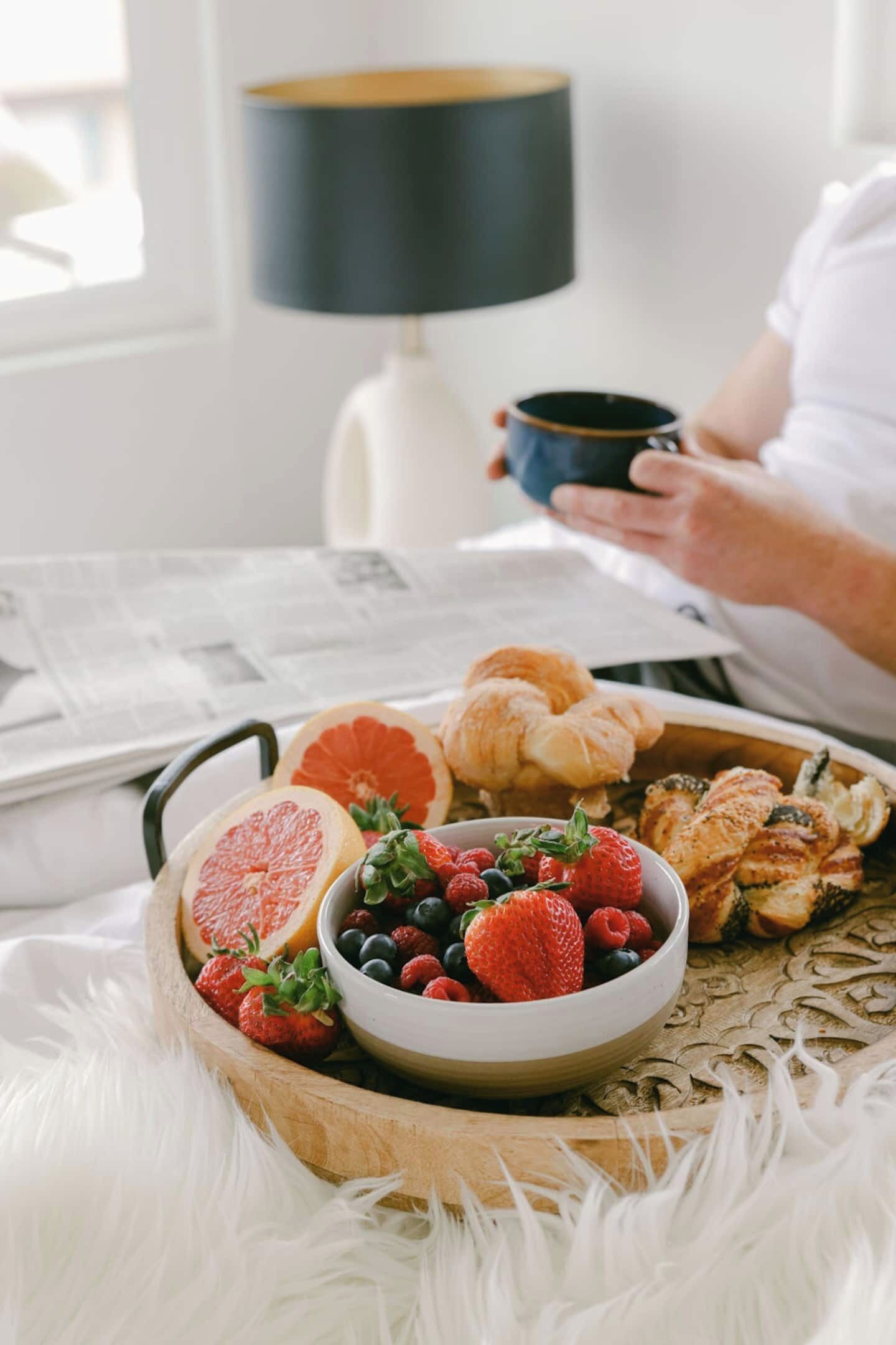 Un plateau de petit-déjeuner avec des fruits et des viennoiseries, une lampe et un journal sur une table.