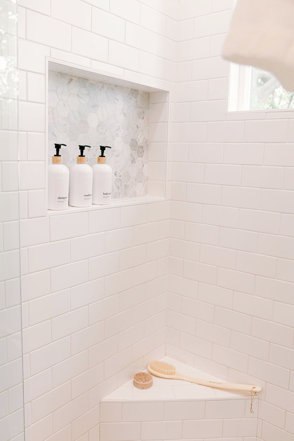 A bathroom with white tiles, a shelf with three bottles of shampoo, a wooden brush, and a soap bar.