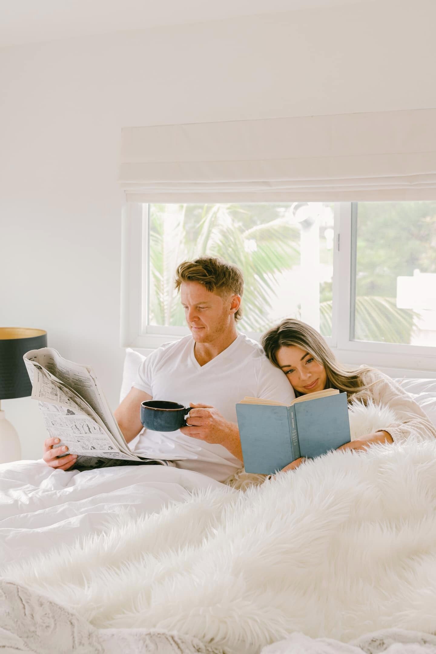 A couple in a bedroom with a large window, a fluffy white bedspread, and a lamp on a bedside table.