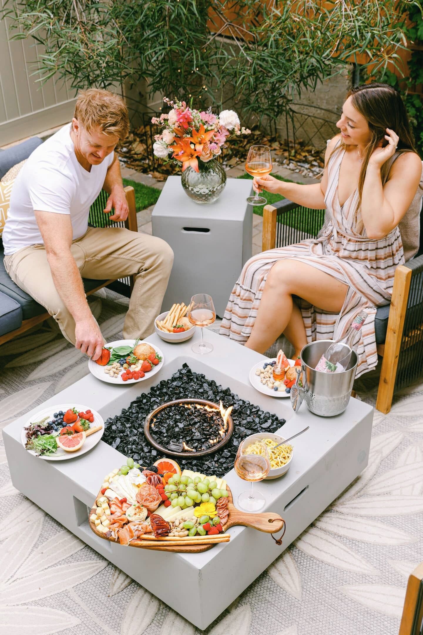 A dining area with a central fire feature, surrounded by a variety of food platters and a bottle of wine.