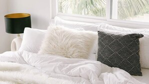 Uma cama com roupa de cama branca e almofada estampada, uma luminária e uma janela com vista para palmeiras.