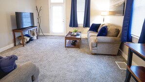 Un salon avec un ventilateur de plafond, un téléviseur posé sur un meuble, un canapé, une table basse et une porte avec des rideaux.