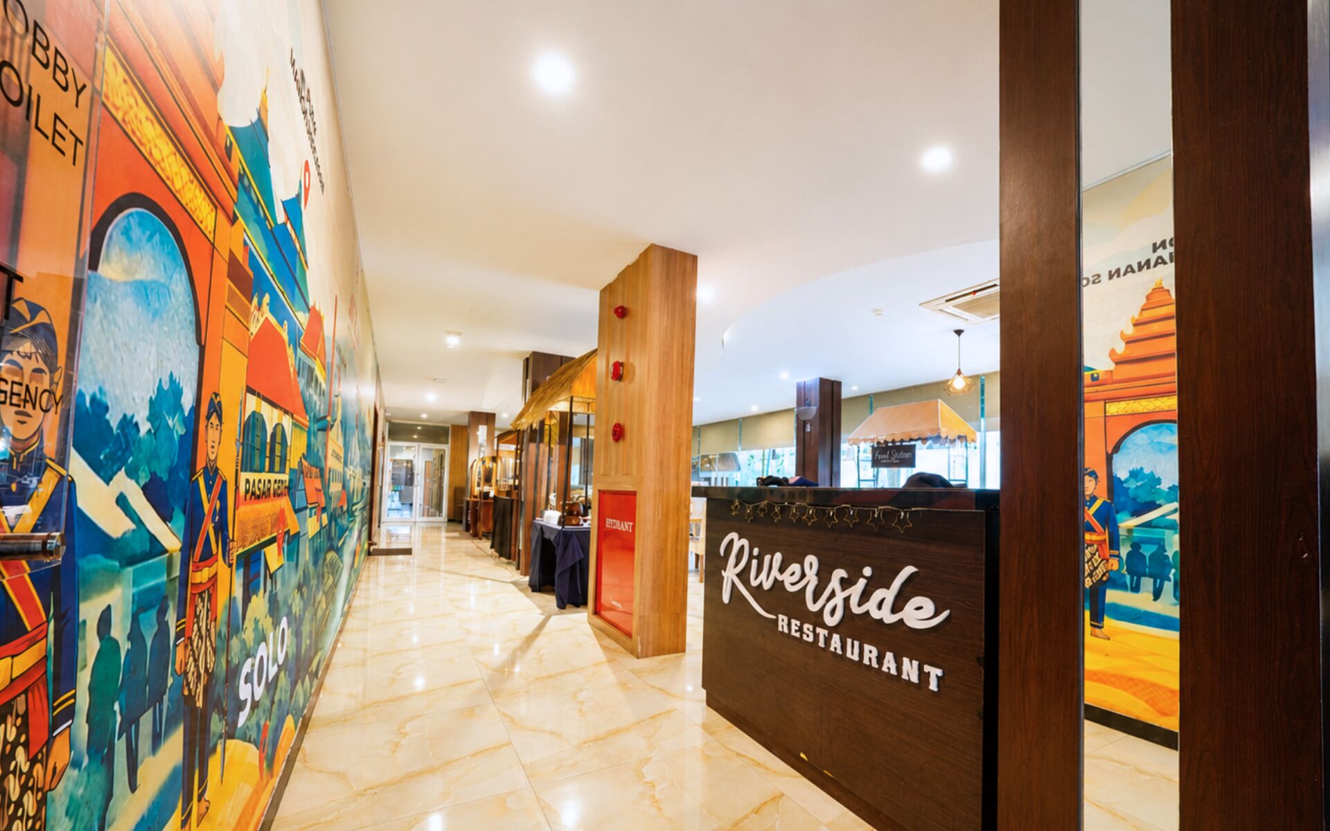 Un pasillo con un mural a la izquierda, un mostrador de recepción de madera con la leyenda 'Riverside Restaurant' y una vista parcial de un restaurante al fondo.