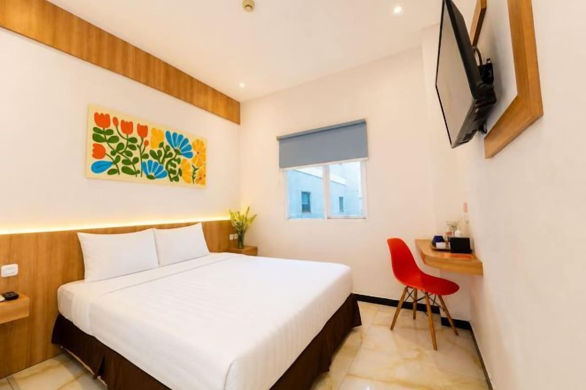 Quarto de hotel com cama, escrivaninha com cadeira, TV fixada na parede e janela com cortina azul.