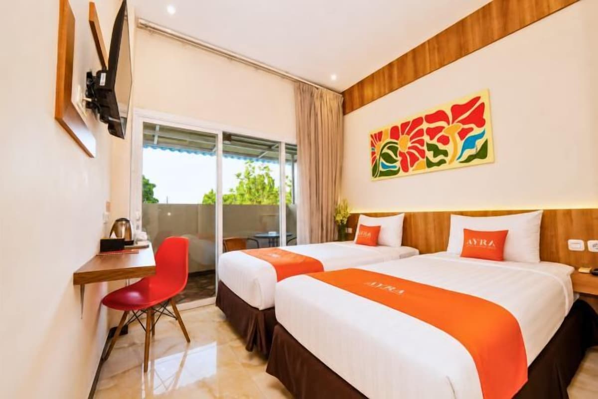 Quarto de hotel com duas camas, uma escrivaninha, uma cadeira vermelha e uma janela grande.