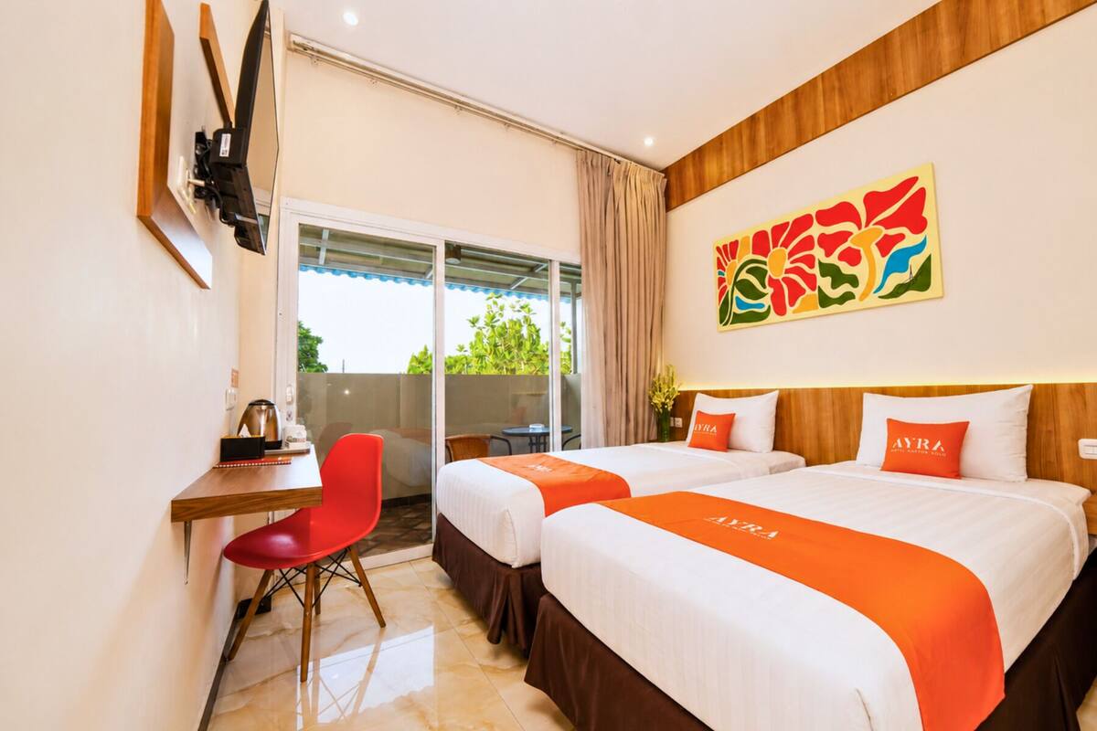 Une chambre d’hôtel avec deux lits, un bureau, une chaise rouge, une porte-fenêtre coulissante donnant sur un balcon et un tableau abstrait aux couleurs vives accroché au mur.
