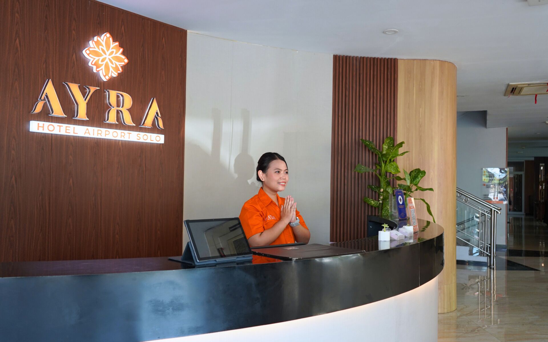 A reception desk with a logo for 'AYRA HOTEL AIRPORT SOLO' and a receptionist in an orange uniform.