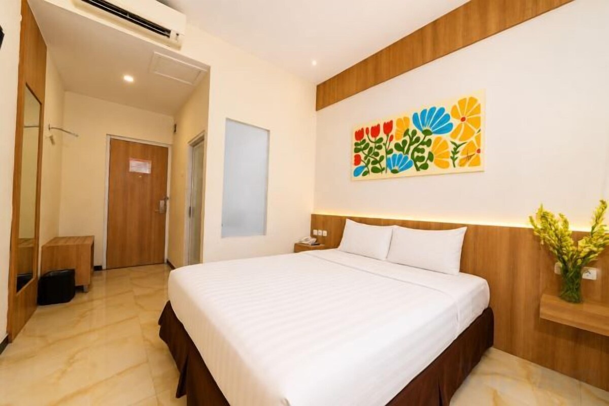 A hotel room with a large bed, a wooden headboard, a nightstand, a bedside table with a lamp, and a wall-mounted artwork.