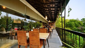 An outdoor dining area with wicker chairs and tables, a covered walkway, and a view of lush greenery. - Ayra Hotel Airport Solo (Ngemplak)