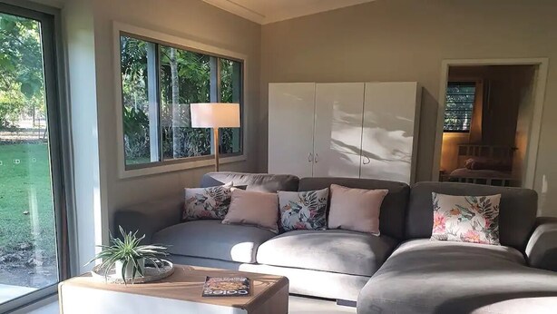 A modern living room with a sectional sofa, a coffee table, and a large window providing a view of the outdoors. - Adorable 2 bedroom unit, set in lush gardens (Mcminns Lagoon)