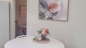A round dining table with four white chairs, a framed floral artwork on the wall, and a small vase with flowers and a candle on the table. - Adorable 2 bedroom unit, set in lush gardens (Mcminns Lagoon)