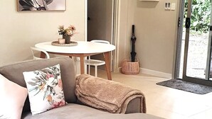 A living room with a sofa, a round table with a vase of flowers, and a door leading outside. - Adorable 2 bedroom unit, set in lush gardens (Mcminns Lagoon)