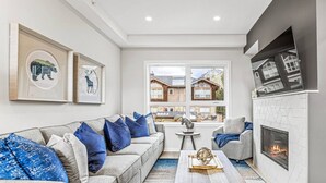 A modern living room with a sofa, coffee table, fireplace, and large windows overlooking a residential area. - Brand New 3BR with Mountain views & Private garage (Canmore)