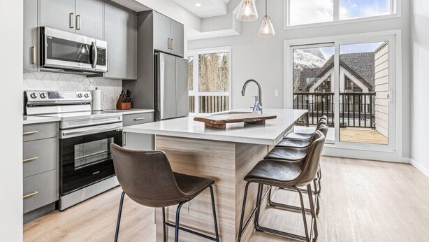 A modern kitchen with a central island, bar stools, and a view of a wooden deck. - Brand New 3BR with Mountain views & Private garage (Canmore)