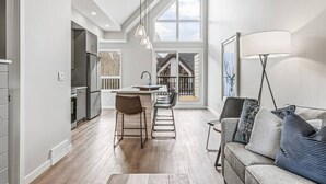 A modern kitchen with a central island, bar stools, and a view of a wooden deck through large windows. - Brand New 3BR with Mountain views & Private garage (Canmore)