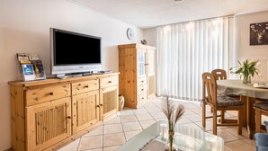 A living room with a TV, wooden cabinet, dining table, chairs, and a vase with plants. - Apartment 'Schwarzwaldblick' with Mountain View, Balcony, and Wi-Fi (St.Georgen im Schwarzwald)