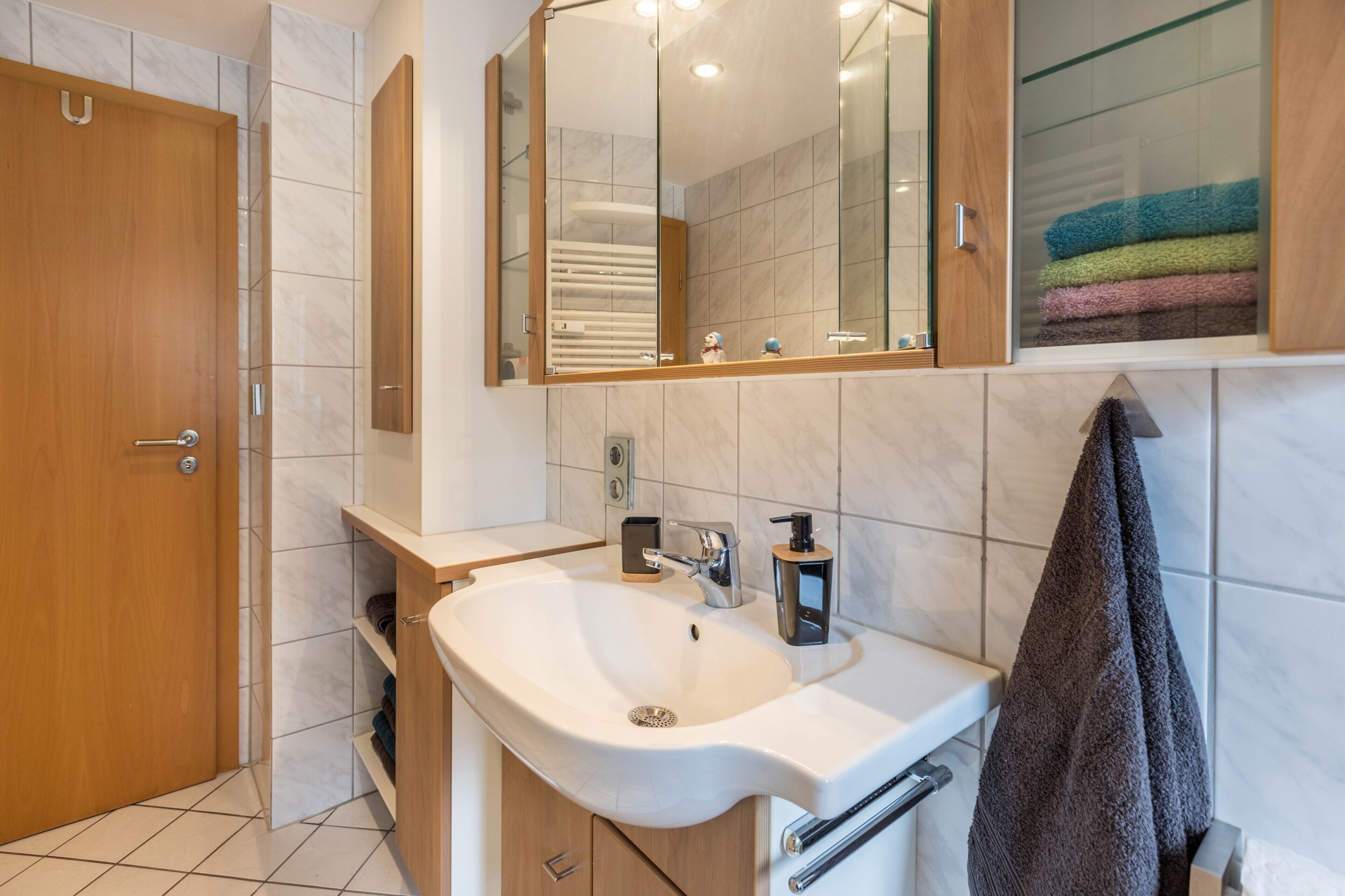Un bany amb una porta de fusta, un lavabo blanc amb un mirall a dalt i un penjallatovelles.